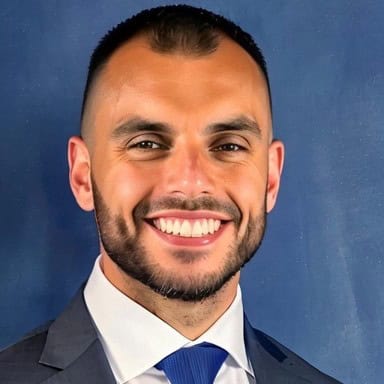 Headshot of a smiling man with short dark hair and a beard, wearing a dark suit, white shirt, and blue tie against a blue background.
