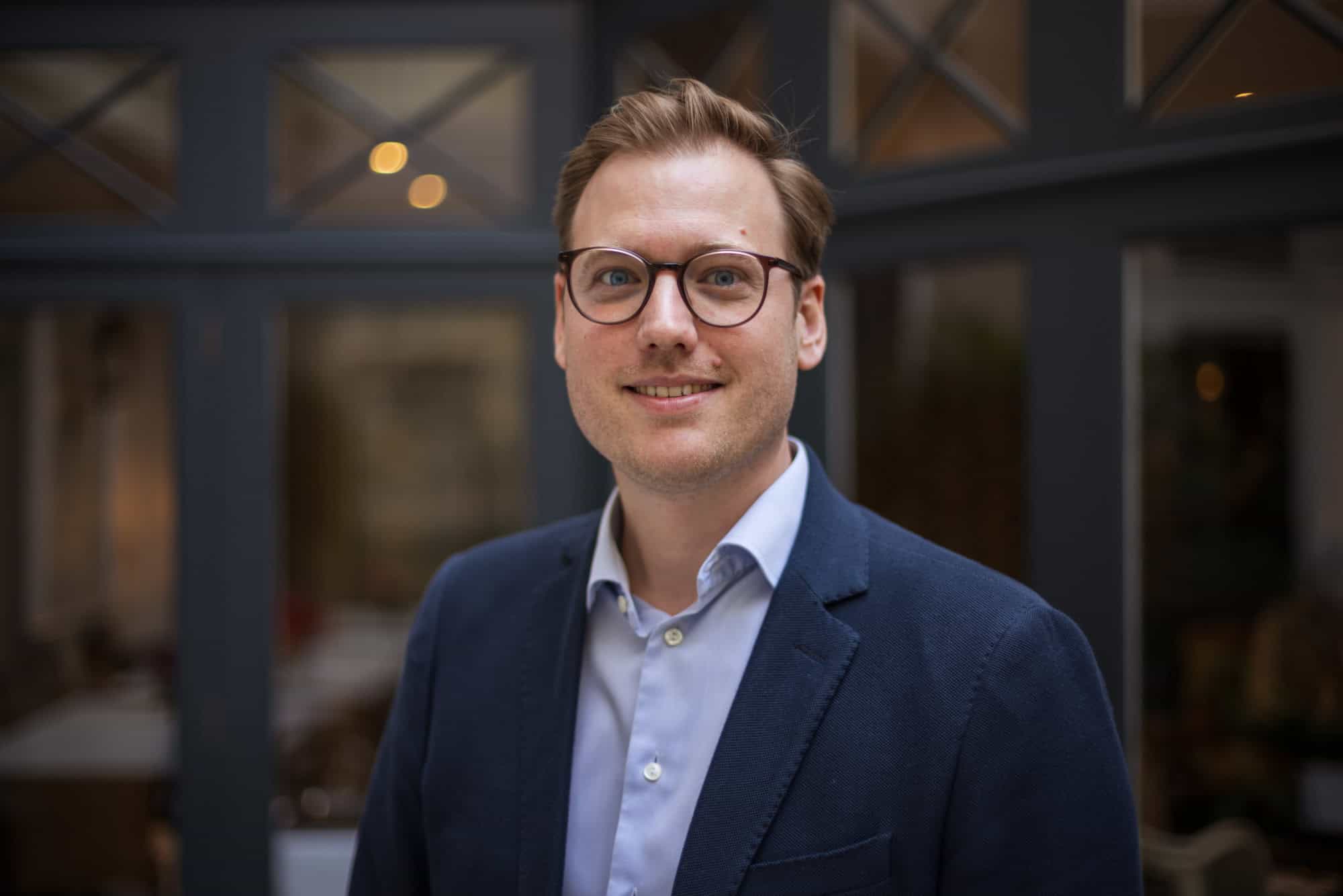 Headshot of a smiling man with light brown hair, blue eyes, and dark-rimmed glasses, wearing a blue shirt and blazer.