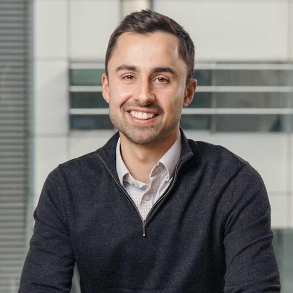 Headshot of a smiling young man with short dark hair and a light beard, wearing a dark gray quarter-zip sweater over a collared shirt.
