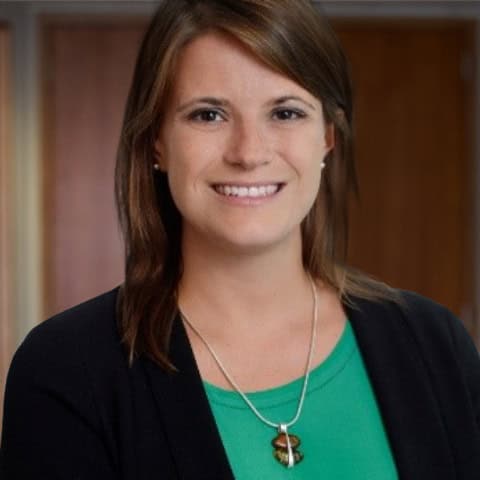 Headshot of a smiling woman with medium brown hair, wearing a green top, black blazer, and a unique amber pendant necklace.