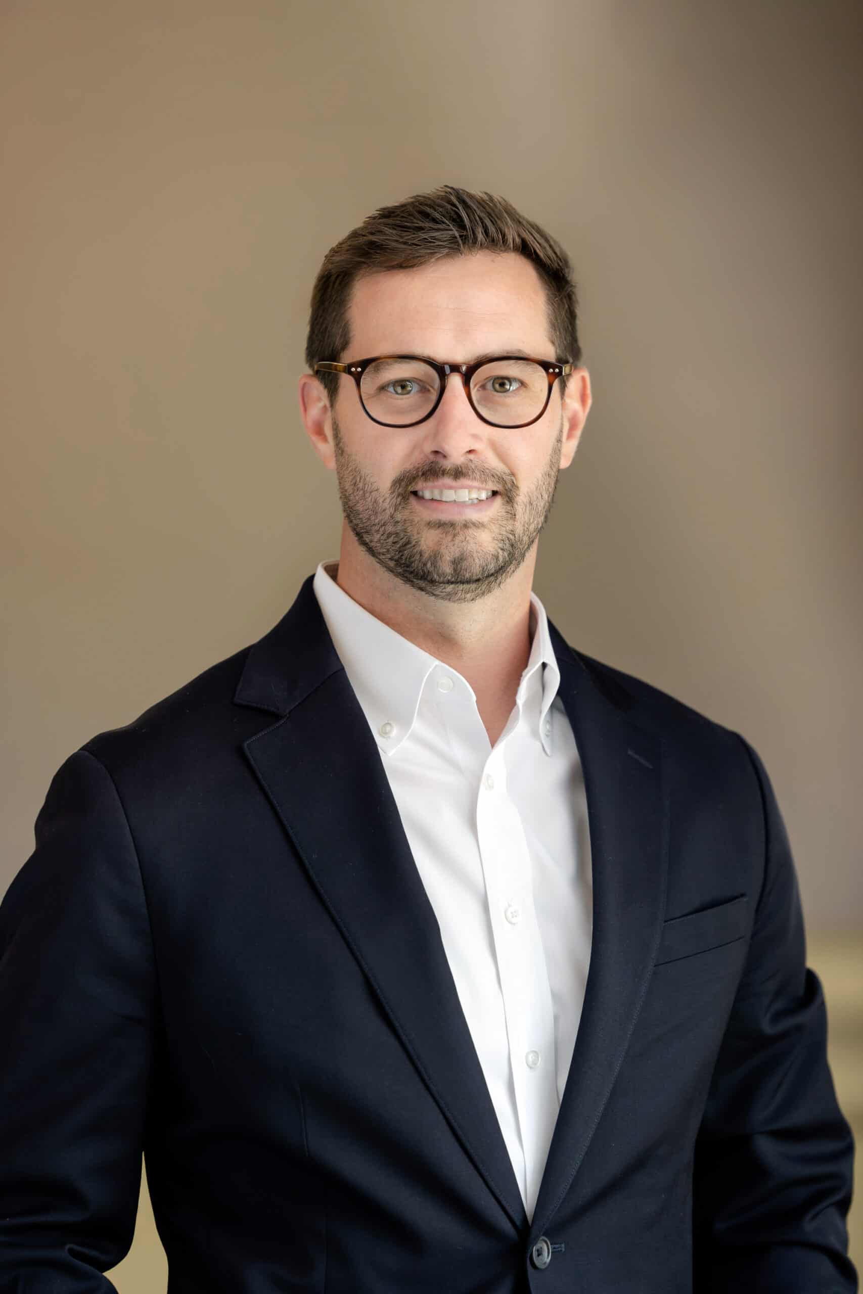Headshot of a man with dark hair, a beard, and glasses, wearing a navy blazer and white shirt, smiling at the camera.