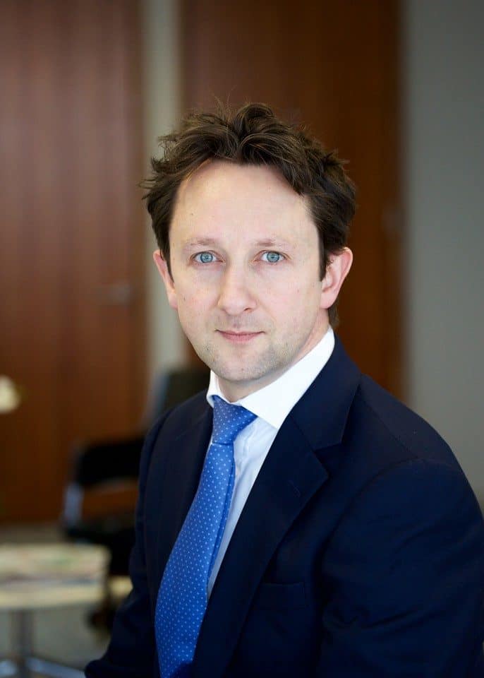 Headshot of a man with blue eyes and dark hair, wearing a blue suit and polka dot tie.