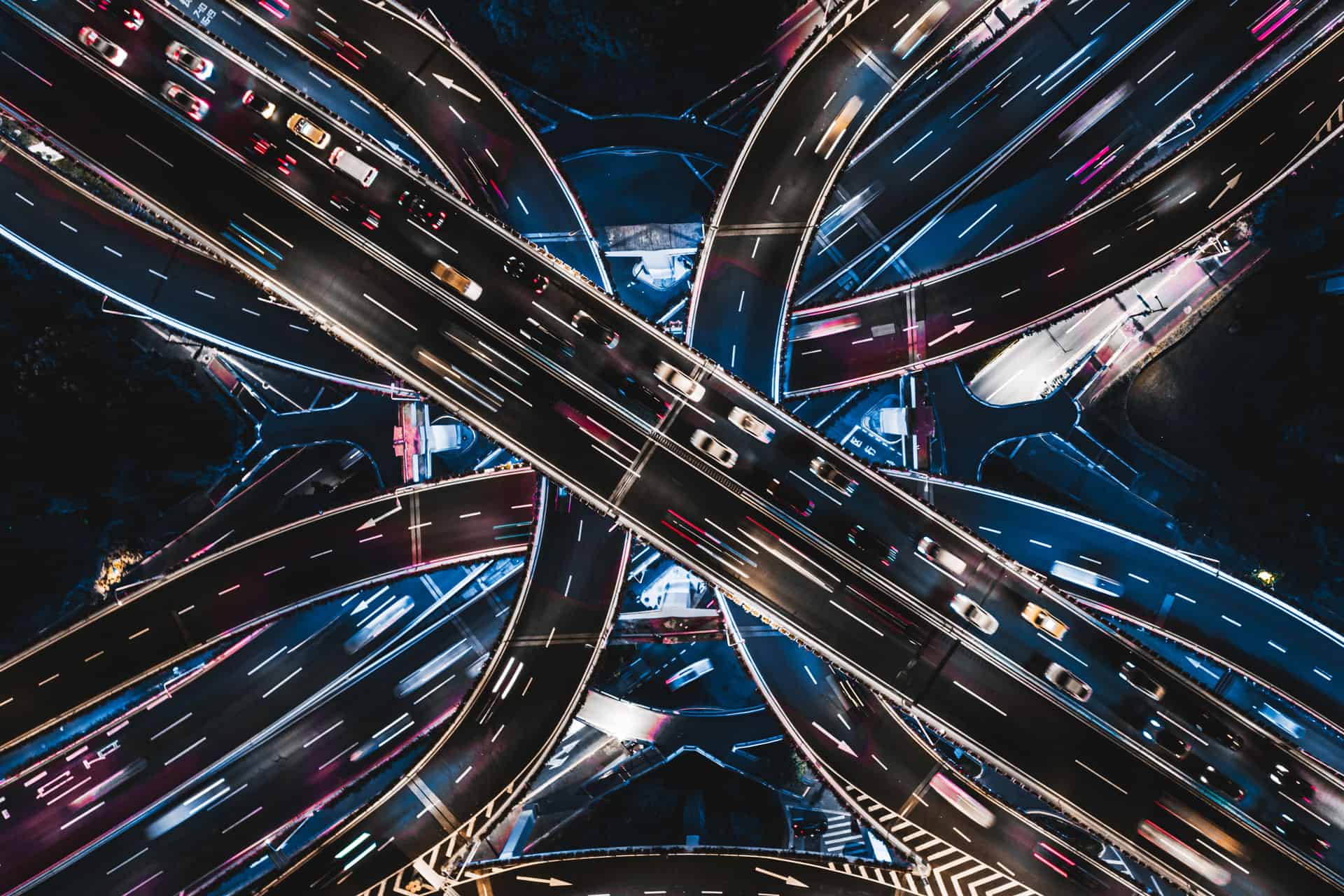 Aerial long-exposure shot of a highway interchange at night, showing vehicles as streaks of white, blue, red, and pink light on dark roads.