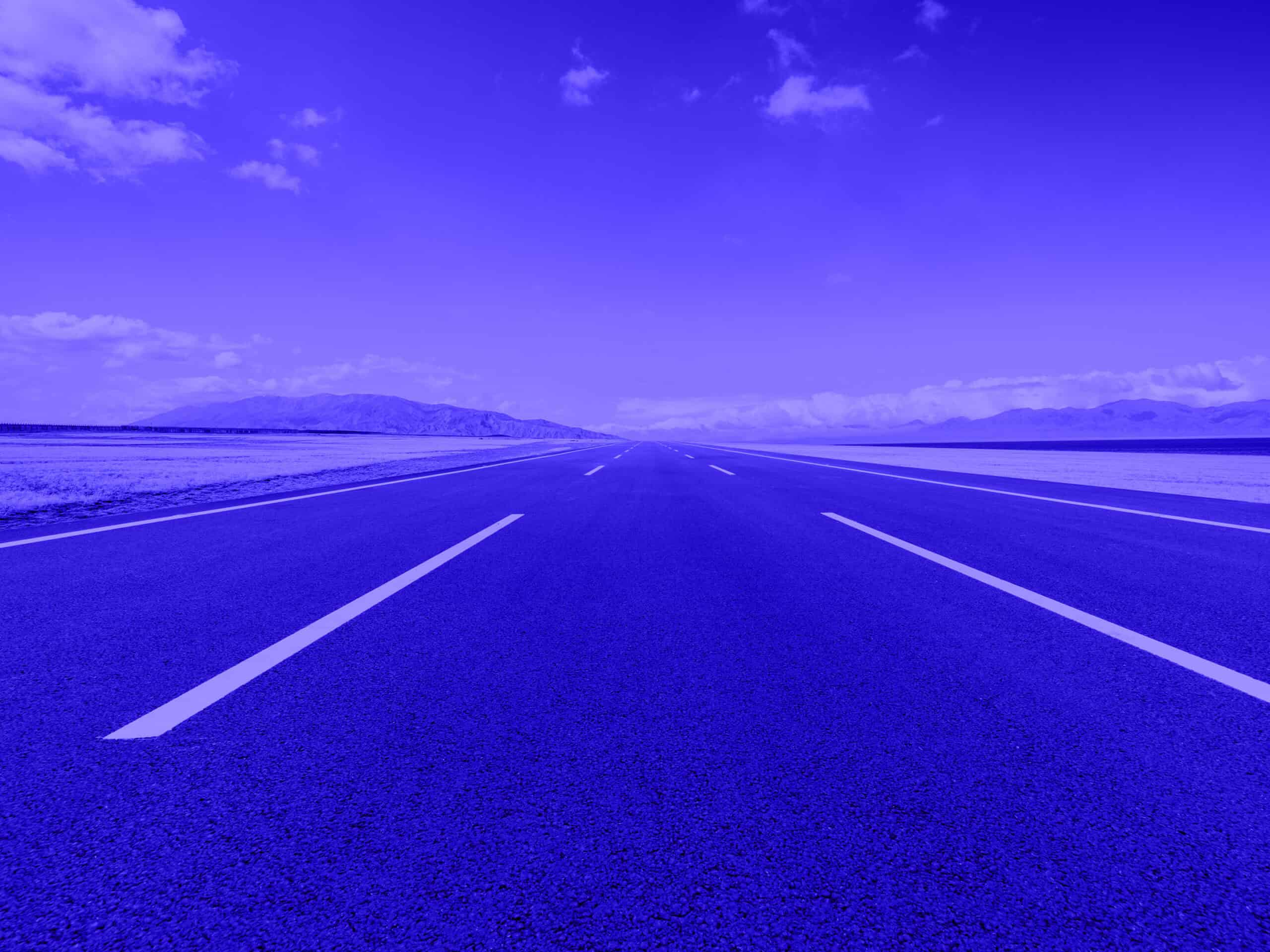 A long, empty blue asphalt road with white markings extends into the distance under a blue sky with white clouds, flanked by blue plains and mountains.
