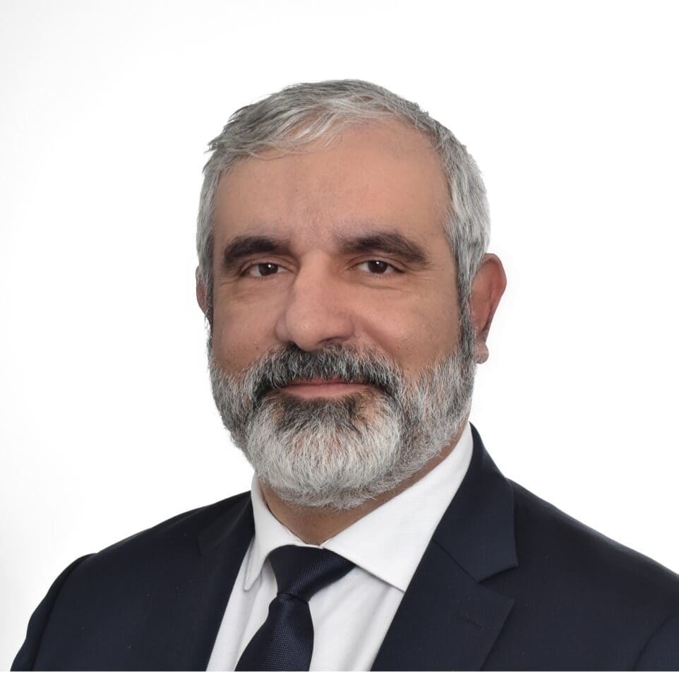 Headshot of a man with gray hair and beard, wearing a navy suit and tie, against a white background.