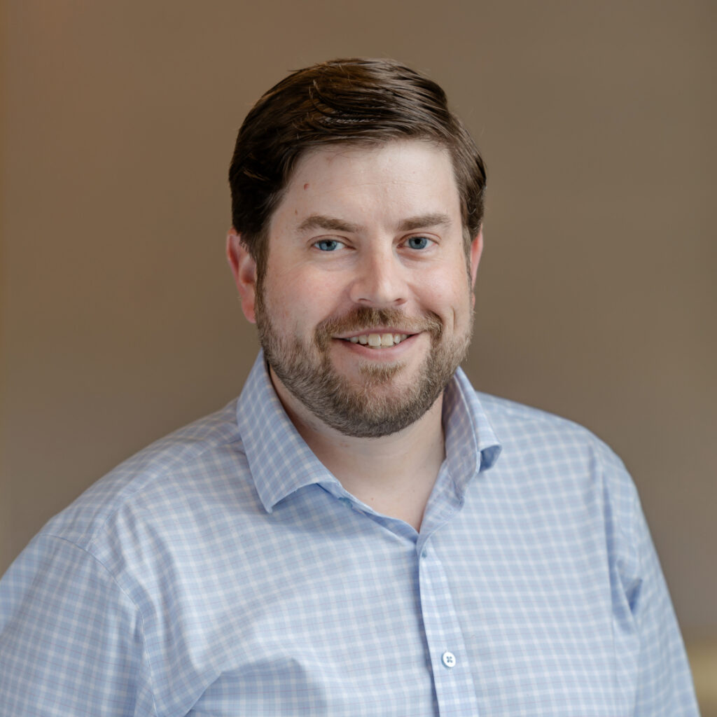 A man with blue eyes and a beard smiles, wearing a blue and white checkered shirt.