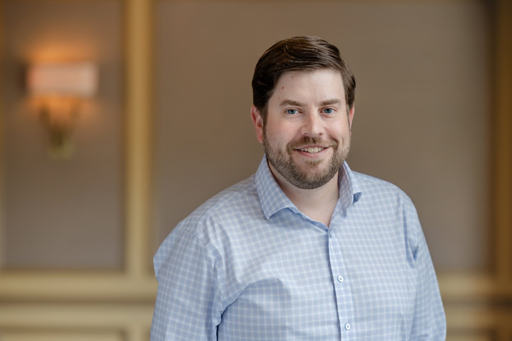 A man with blue eyes and a beard smiles, wearing a blue and white checkered shirt.