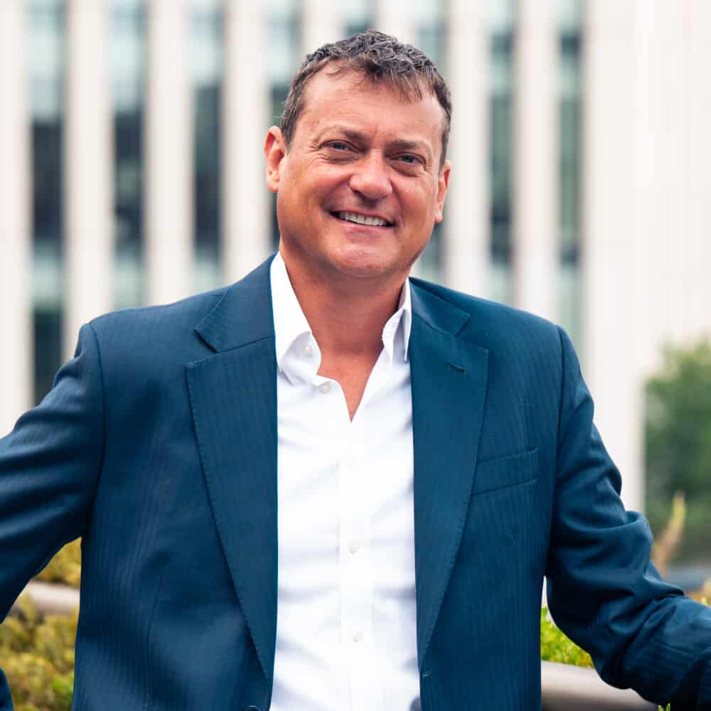A smiling man in a dark blue suit and white shirt stands outdoors with green plants and modern buildings in the background.