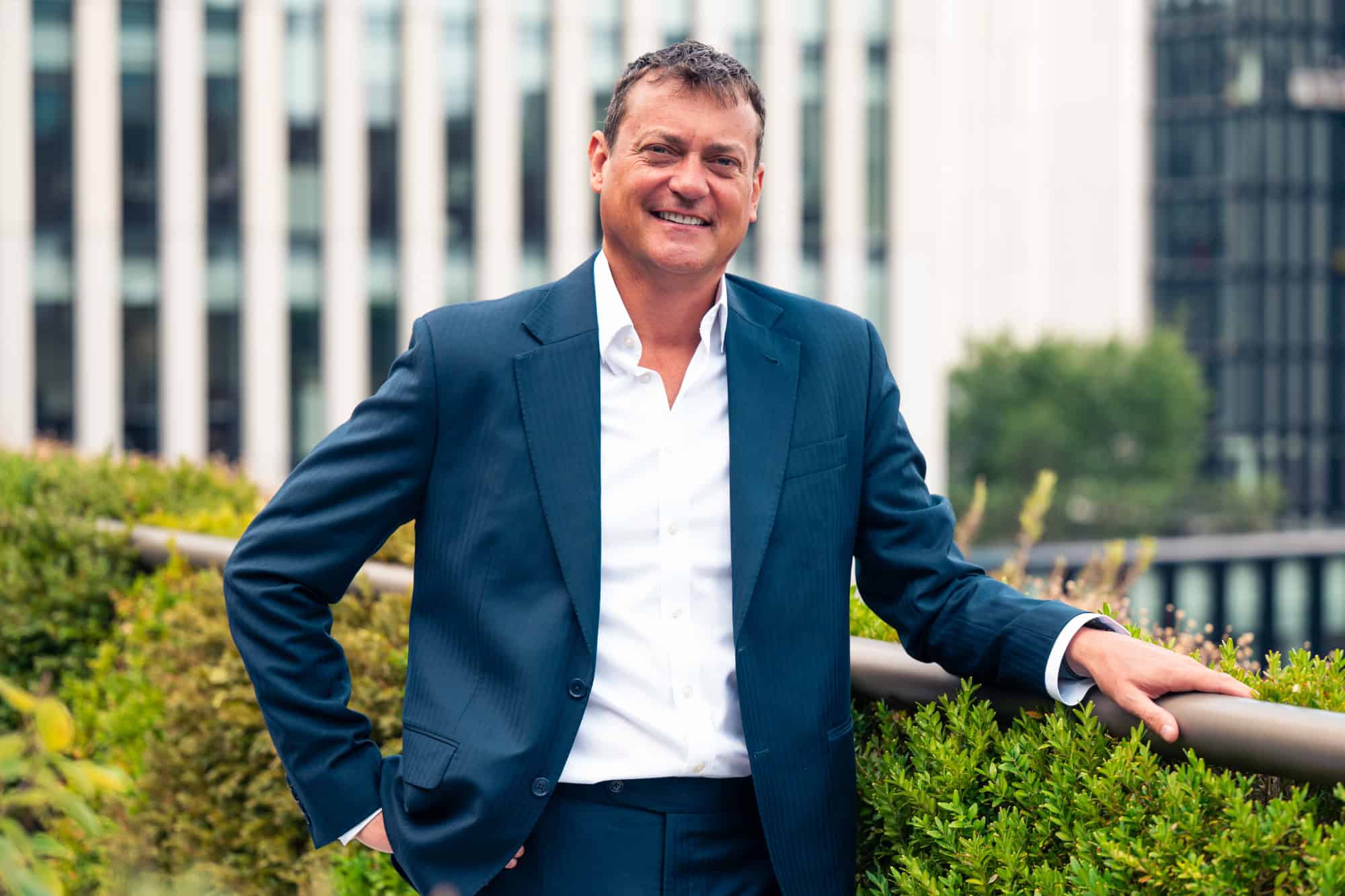 A smiling man in a dark blue suit and white shirt stands outdoors with green plants and modern buildings in the background.