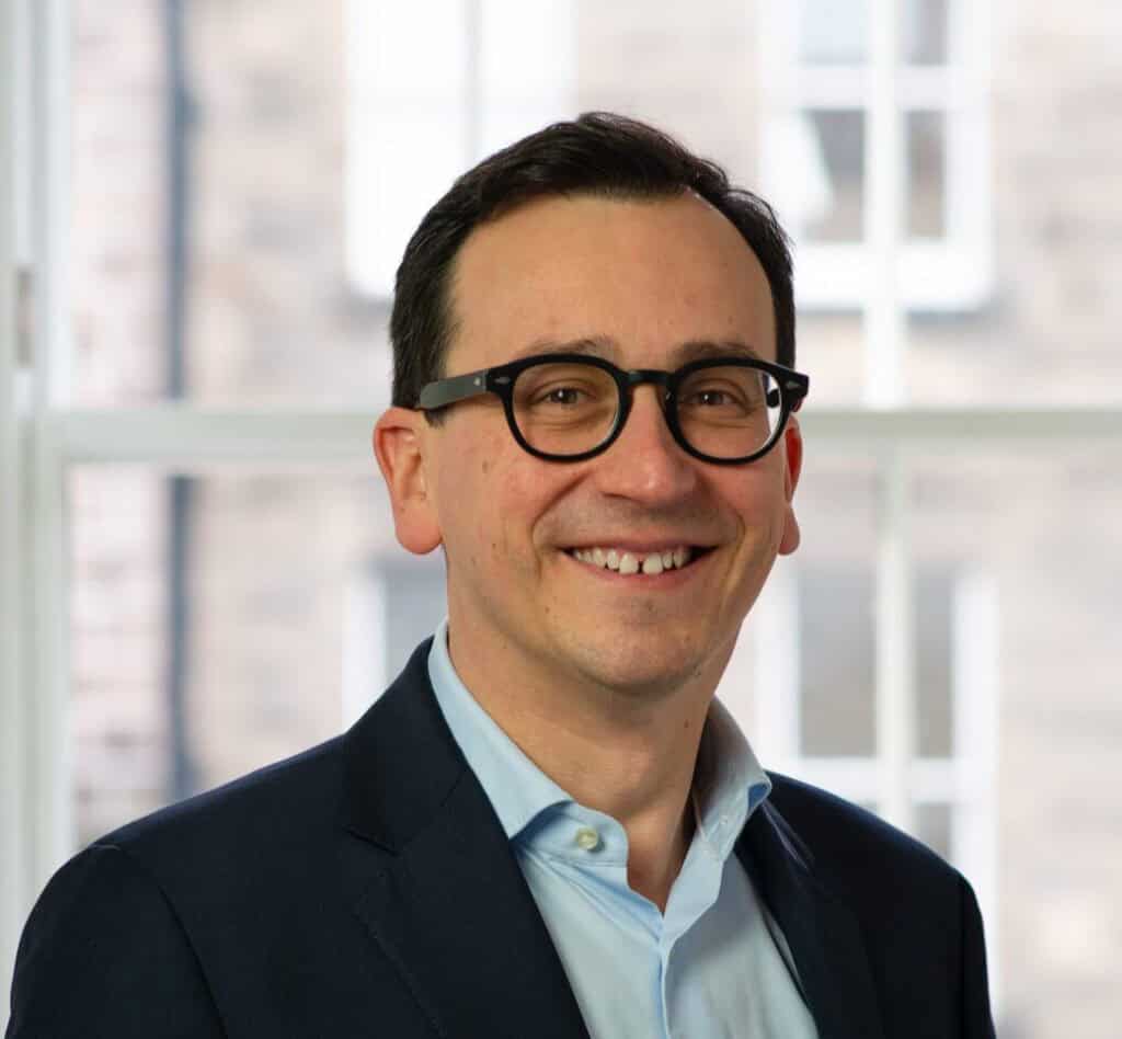 Headshot of a smiling man with dark hair and glasses, wearing a blue shirt and blazer.