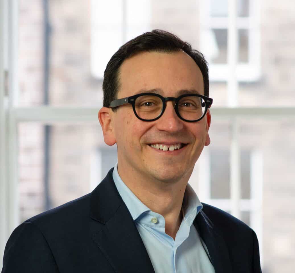 Headshot of a smiling man with dark hair and glasses, wearing a blue shirt and blazer.