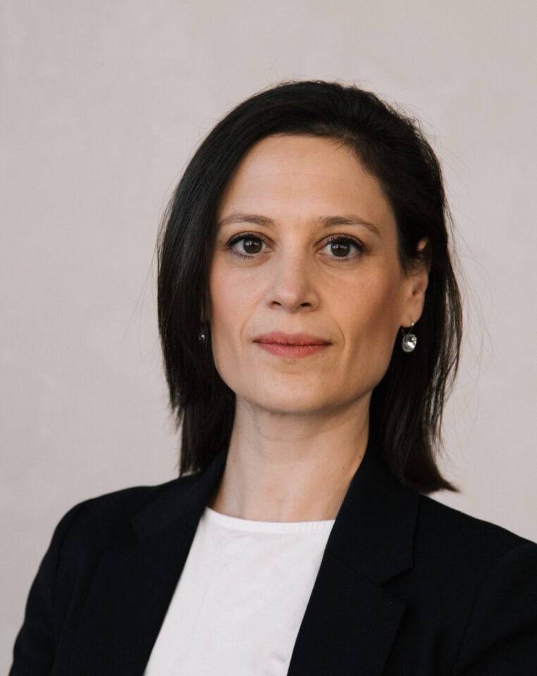 Headshot of a woman with dark hair, light brown eyes, and a neutral expression, wearing a dark blazer over a white top.