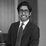 Black and white headshot of a smiling man with dark hair, glasses, and a suit.