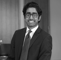Black and white headshot of a smiling man with dark hair, glasses, and a suit.