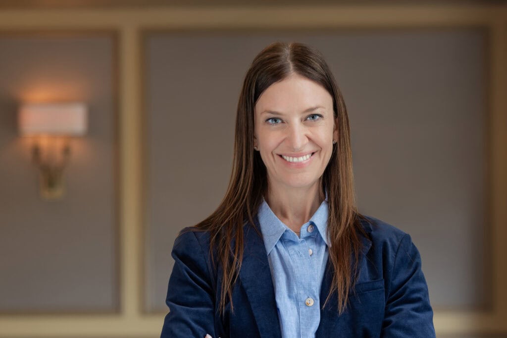 Headshot of a smiling woman with long brown hair and blue eyes, wearing a blue shirt and blazer.