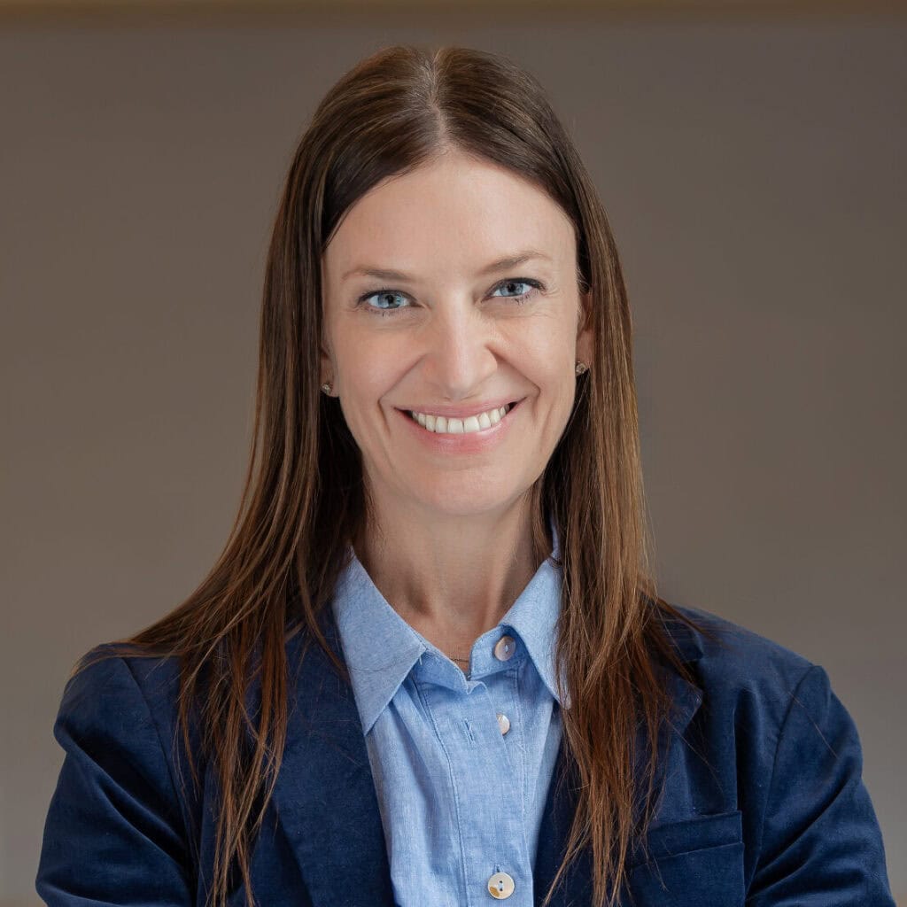 Headshot of a smiling woman with long brown hair and blue eyes, wearing a blue shirt and blazer.