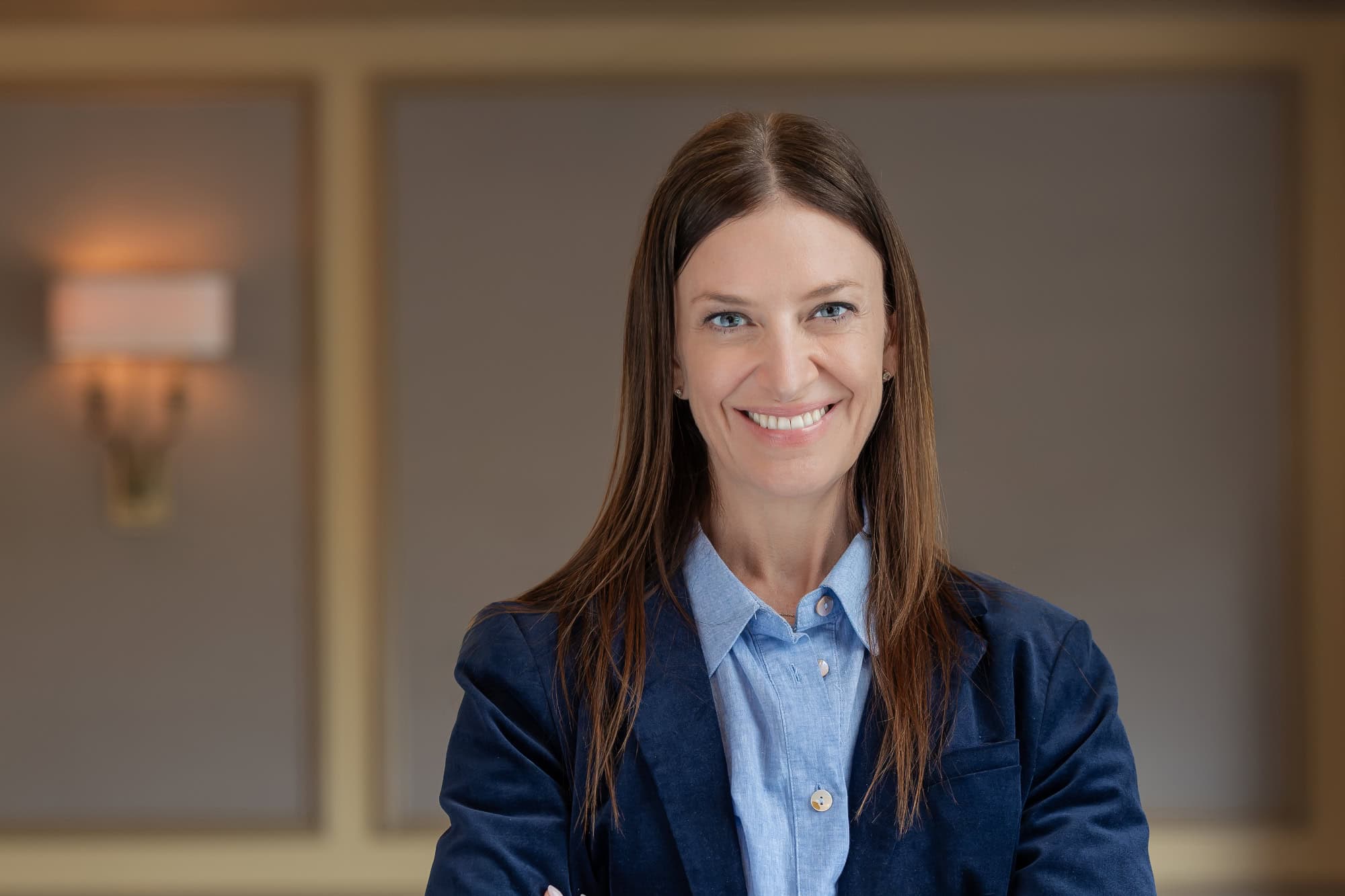 Headshot of a smiling woman with long brown hair and blue eyes, wearing a blue shirt and blazer.