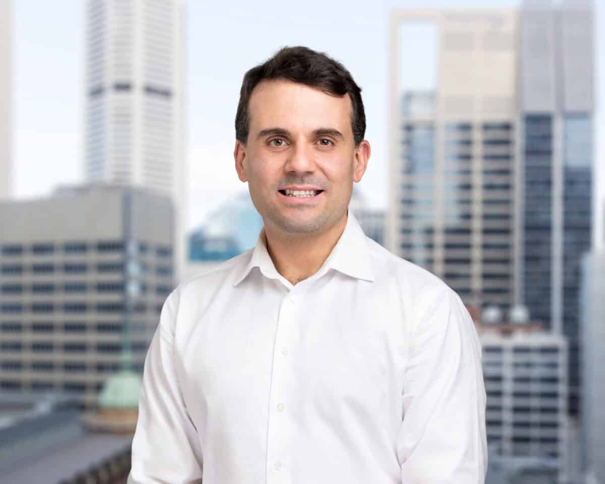 A man with dark hair and a white shirt smiles at the camera, with a blurred city skyline in the background.