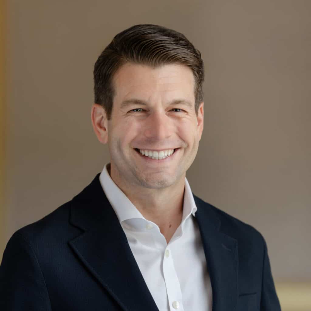 Headshot of a man smiling in a dark suit jacket and white shirt against a blurred indoor background.