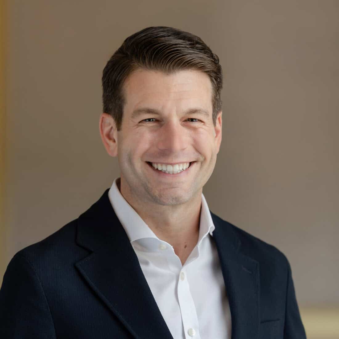 Headshot of a man smiling in a dark suit jacket and white shirt against a blurred indoor background.