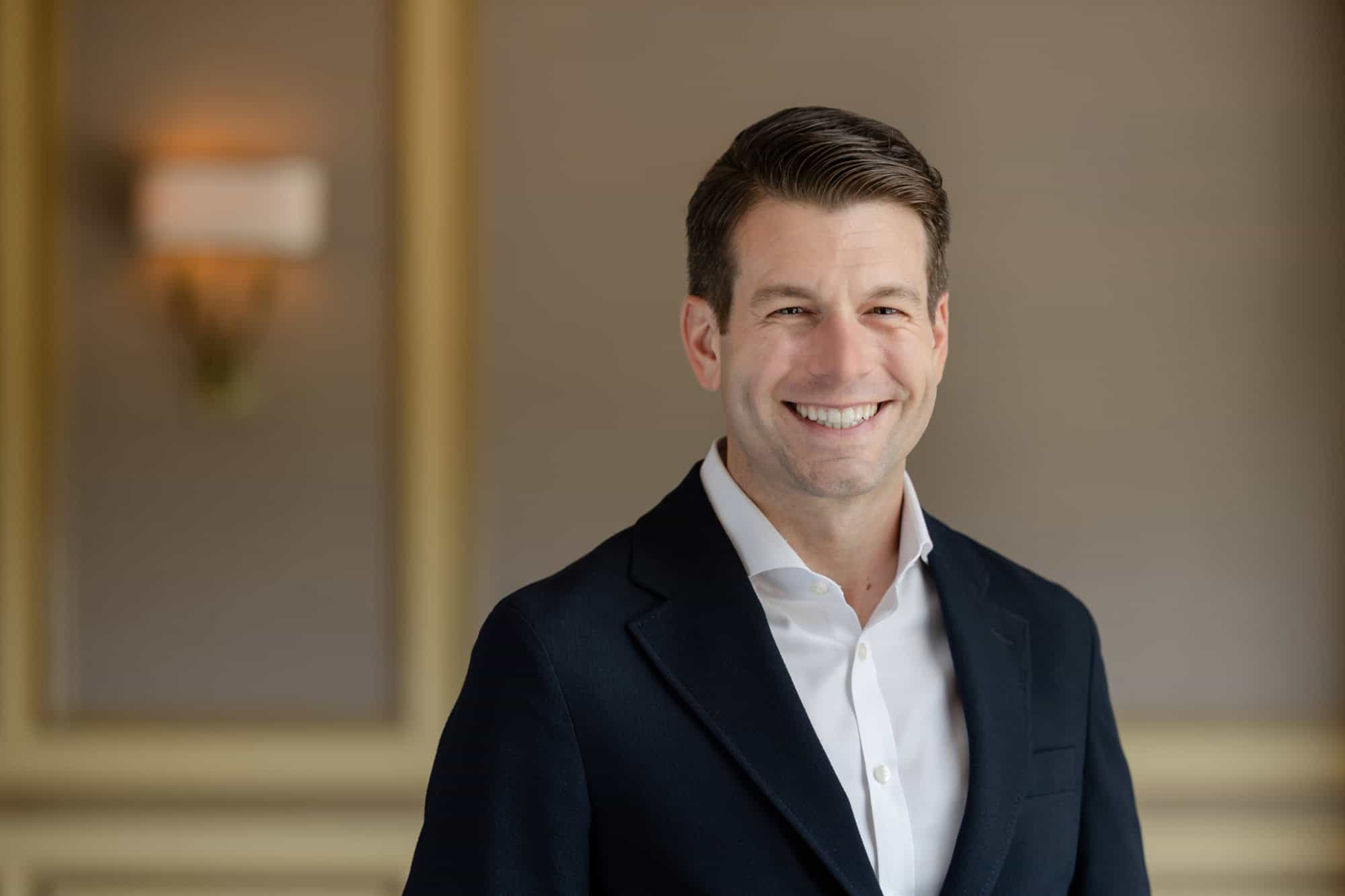 Headshot of a man smiling in a dark suit jacket and white shirt against a blurred indoor background.
