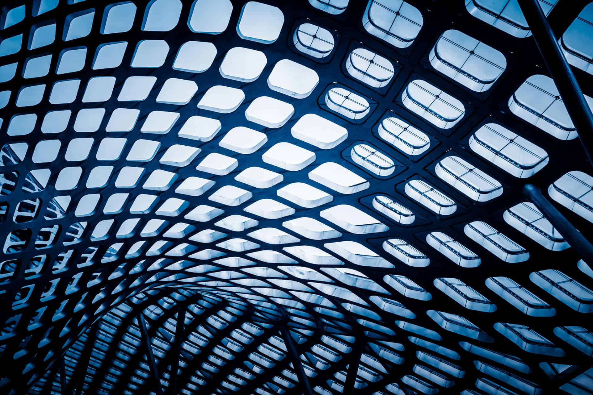 Upward view of a modern ceiling with a dark, intricate grid pattern and numerous blue-tinted skylights.