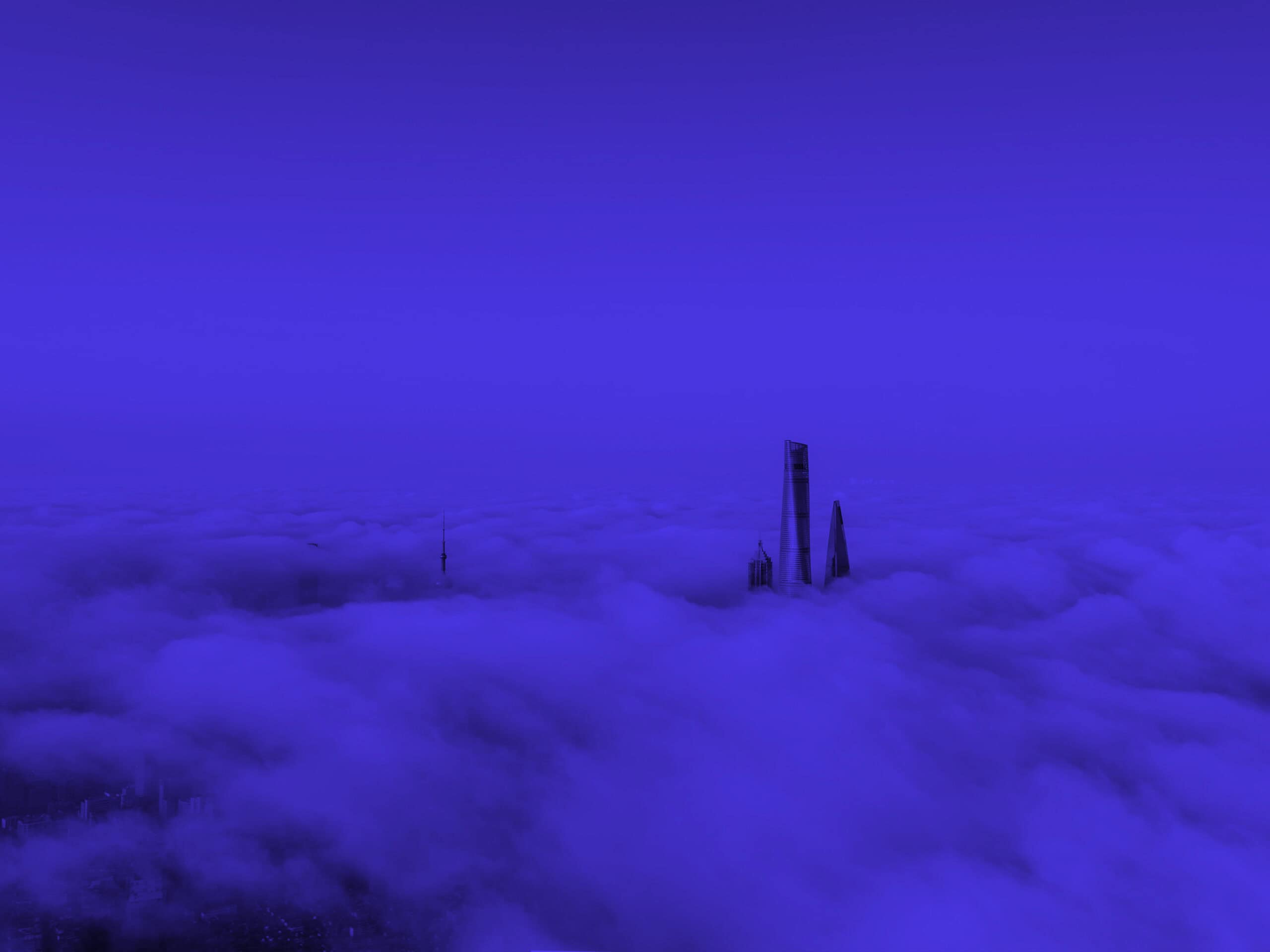 Three tall skyscrapers and a communication tower rise above a dense layer of blue-tinted clouds, with a dark blue sky above.
