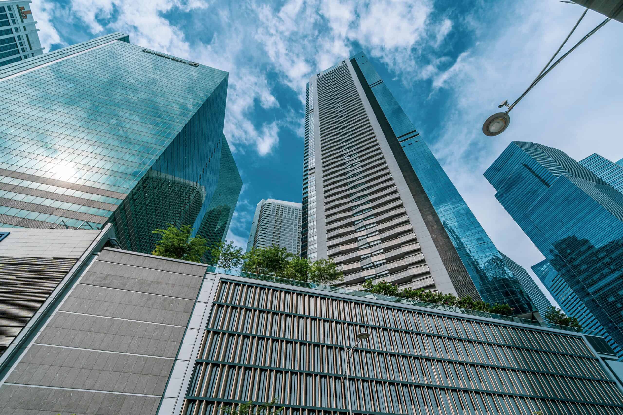 Low-angle view of multiple modern skyscrapers with glass and textured facades under a blue sky with clouds. A streetlight is visible on the right.