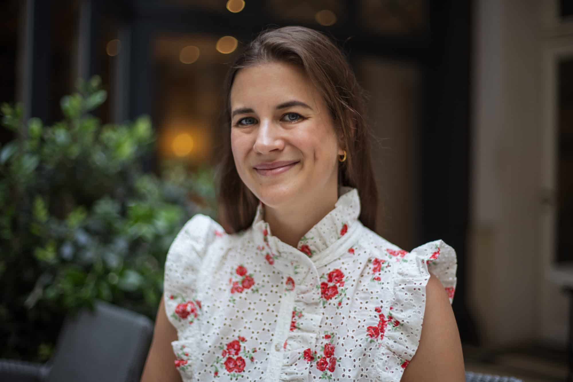 A young woman with brown hair and blue eyes smiles, wearing a white floral top with red roses.