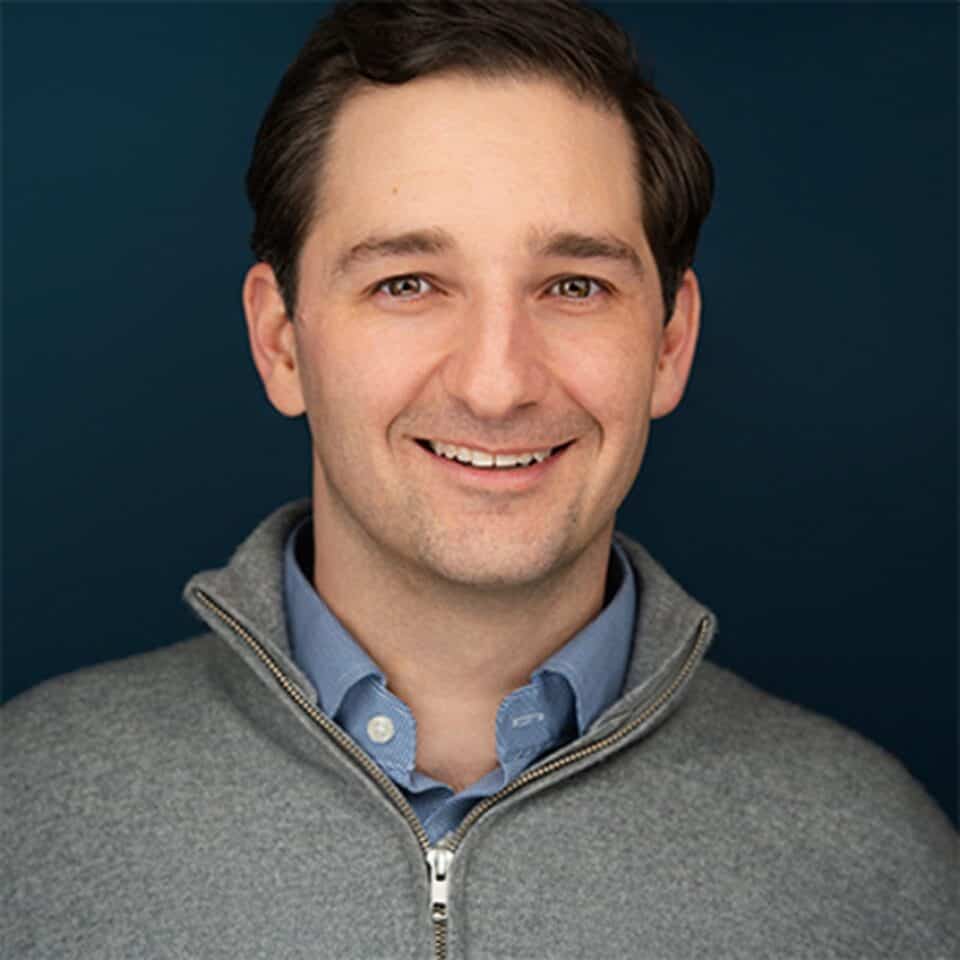 Headshot of a smiling man with dark hair in a gray zippered sweater and blue shirt against a dark blue background.