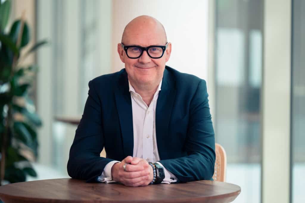 A smiling bald man in a dark suit and black-framed glasses sits at a wooden table with his hands clasped.