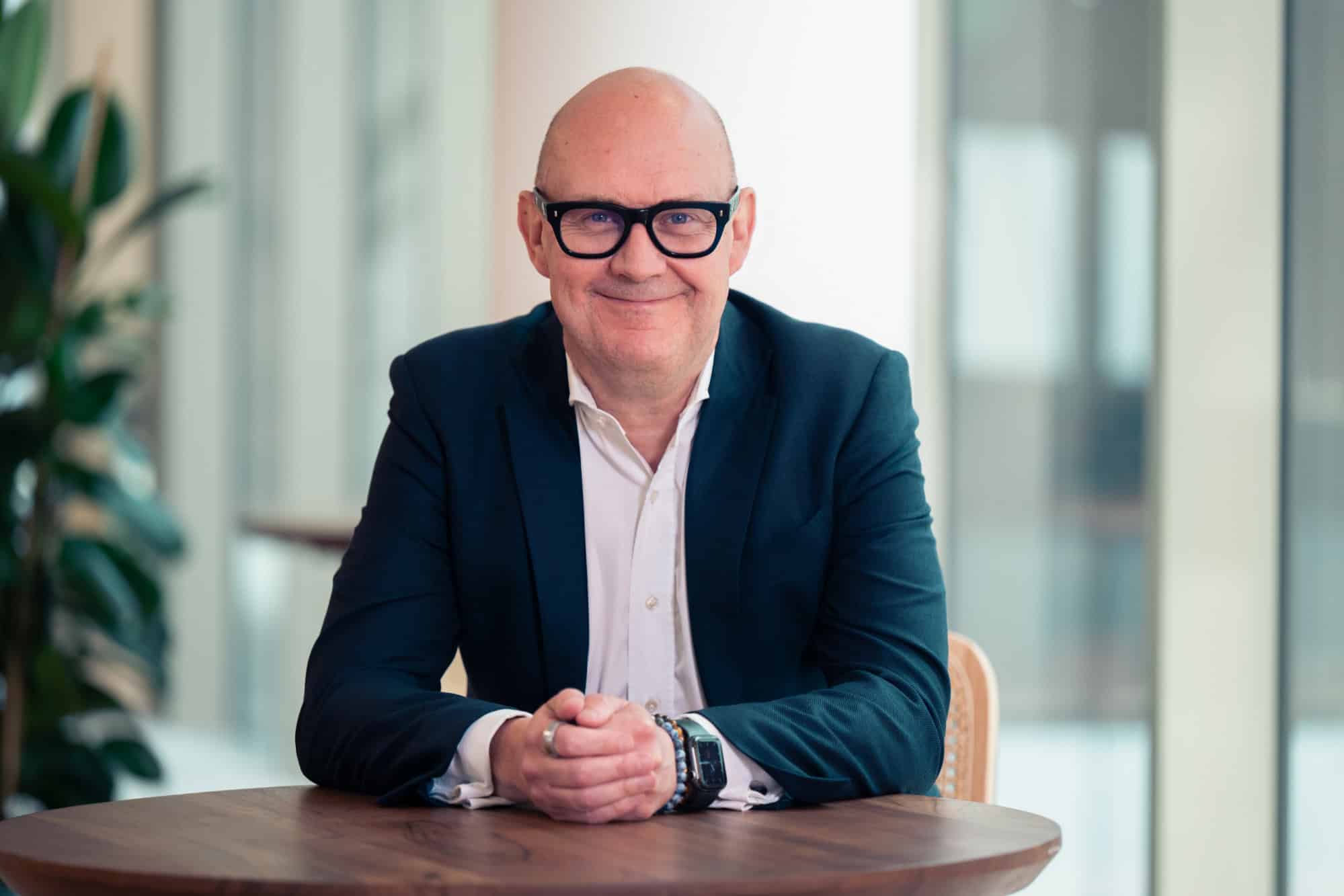 A smiling bald man in a dark suit and black-framed glasses sits at a wooden table with his hands clasped.