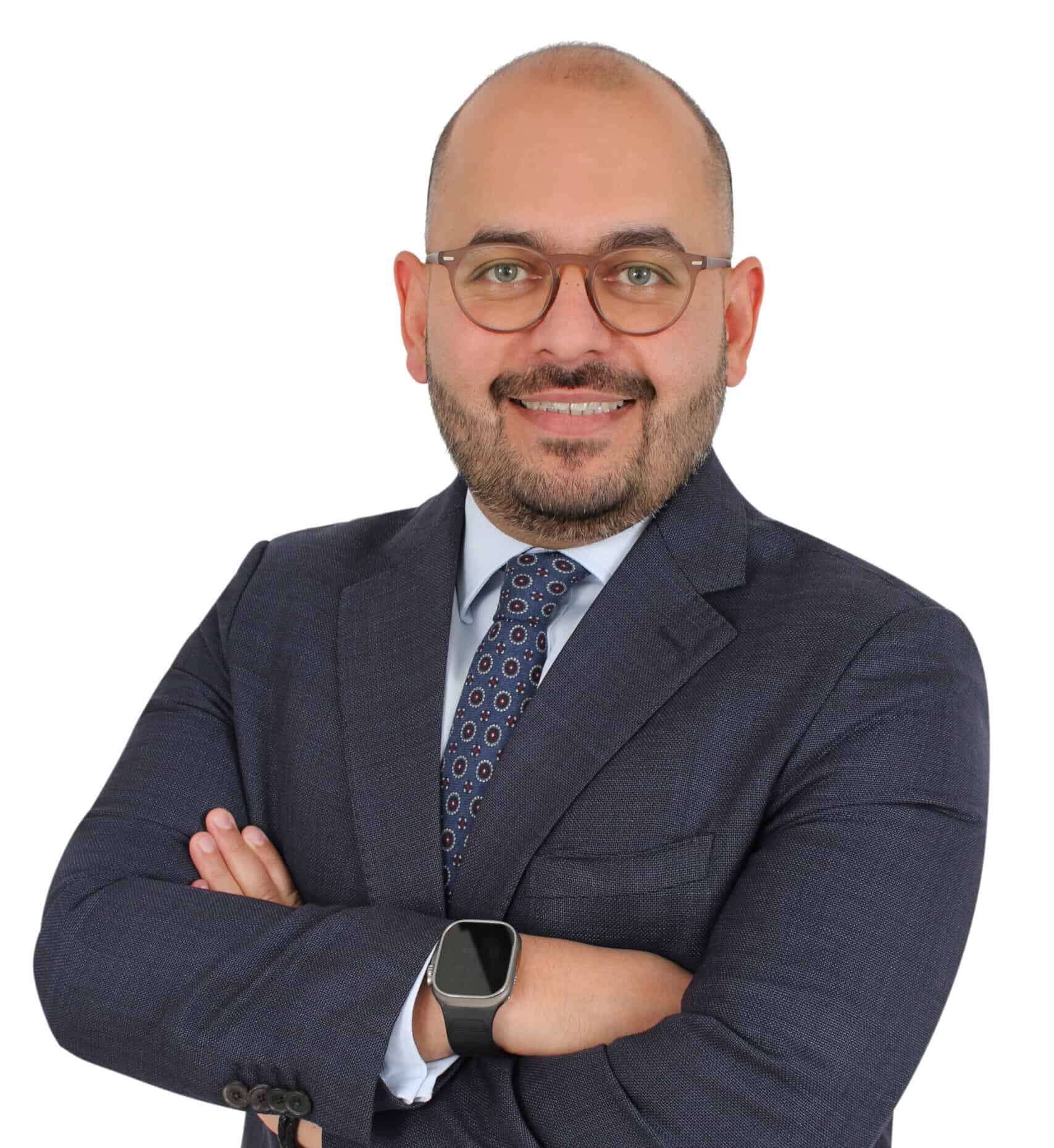 Headshot of a smiling man with a bald head, beard, and glasses, wearing a dark suit and tie, with arms crossed.