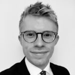 Black and white headshot of a young man with spiky hair, wearing round glasses, a suit, and a tie, smiling slightly.