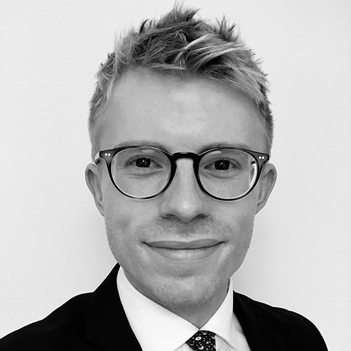 Black and white headshot of a young man with spiky hair, wearing round glasses, a suit, and a tie, smiling slightly.