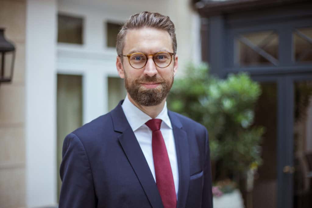 A man with a beard and glasses, wearing a dark suit and red tie, smiles at the camera.