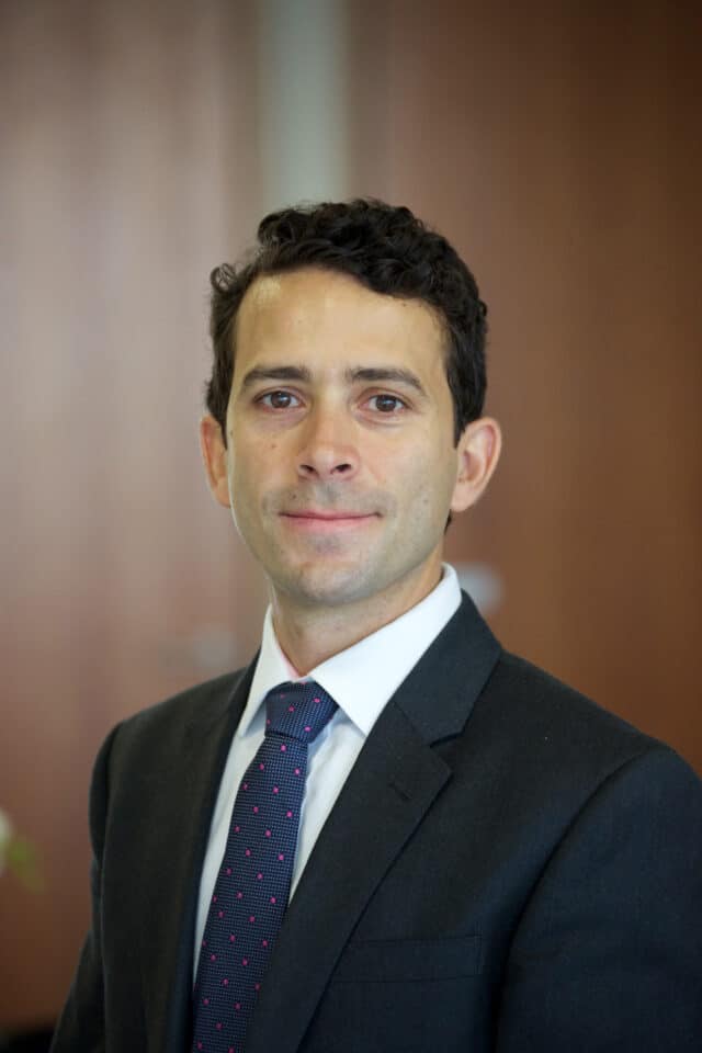Headshot of a man in a dark suit, white shirt, and blue tie with pink polka dots.
