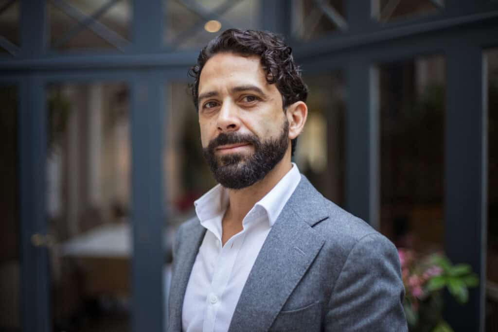 Headshot of a man with dark curly hair and a beard, wearing a gray blazer and white shirt.