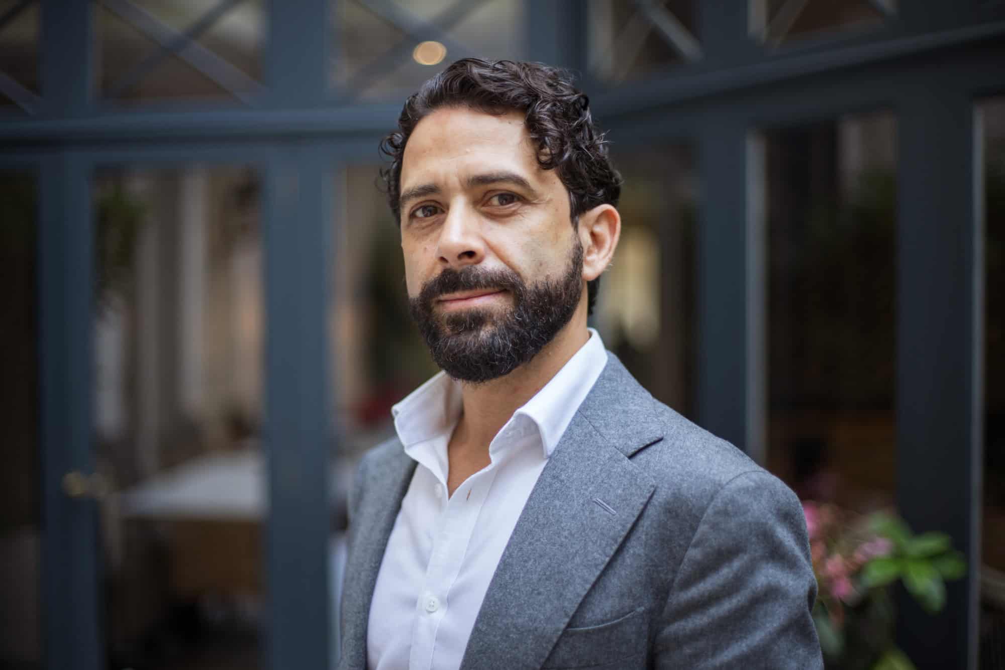 Headshot of a man with dark curly hair and a beard, wearing a gray blazer and white shirt.