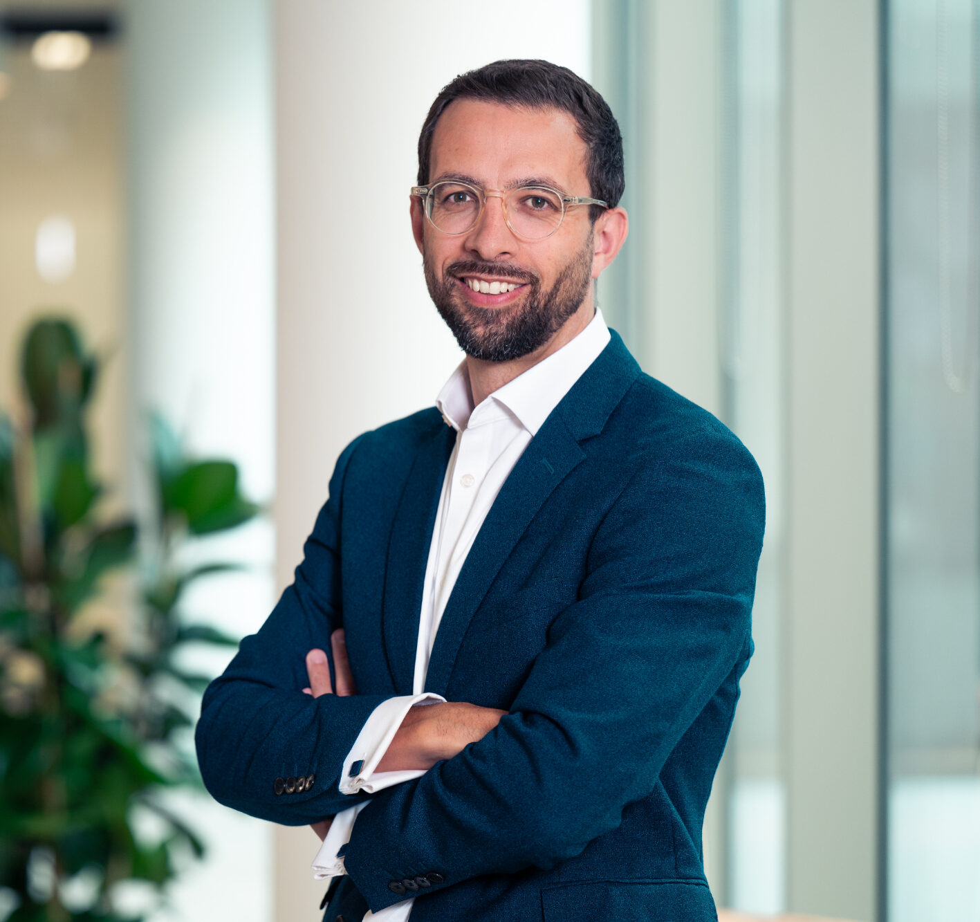 A smiling man with glasses and a beard, wearing a dark teal blazer, stands with his arms crossed in an office setting.