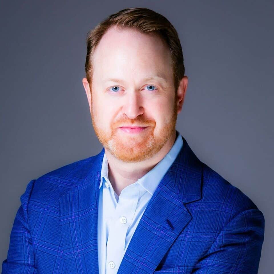 Headshot of a man with reddish-brown hair and beard, blue eyes, wearing a blue plaid suit jacket and light blue shirt, smiling.