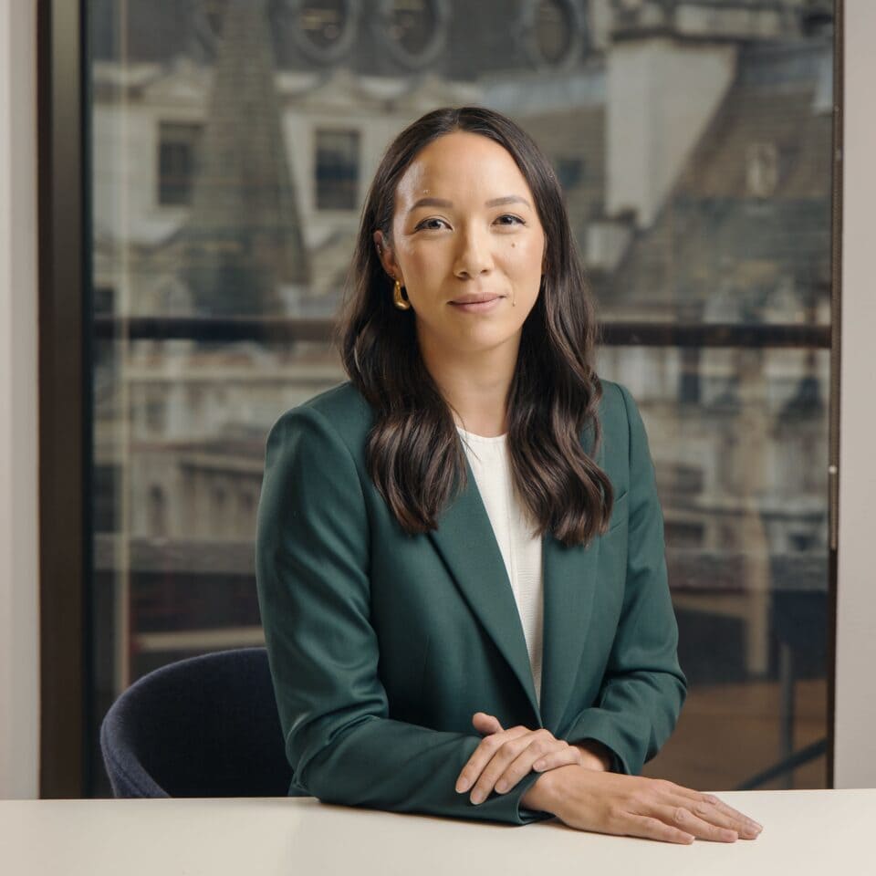 A woman with dark hair and a green blazer smiles while sitting at a table with a blurred city background.