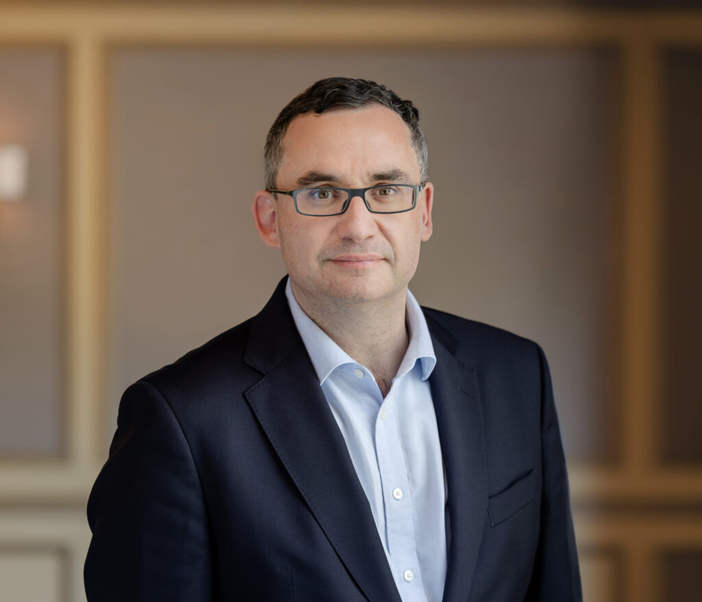 Headshot of a man with short dark hair and glasses, wearing a blue shirt and dark suit jacket, against a blurred background.