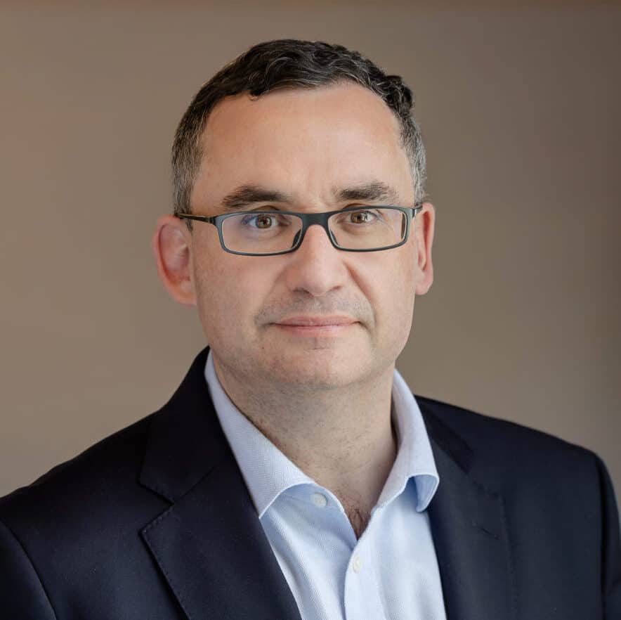 Headshot of a man with short dark hair and glasses, wearing a blue shirt and dark suit jacket, against a blurred background.