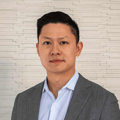 Headshot of an East Asian man in a gray suit jacket and light blue shirt against a textured light-colored wall.