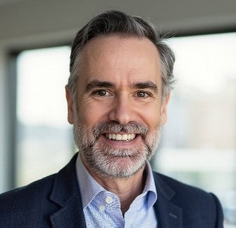 Headshot of a smiling middle-aged man with dark, graying hair and a gray beard, wearing a dark suit jacket and light blue shirt.