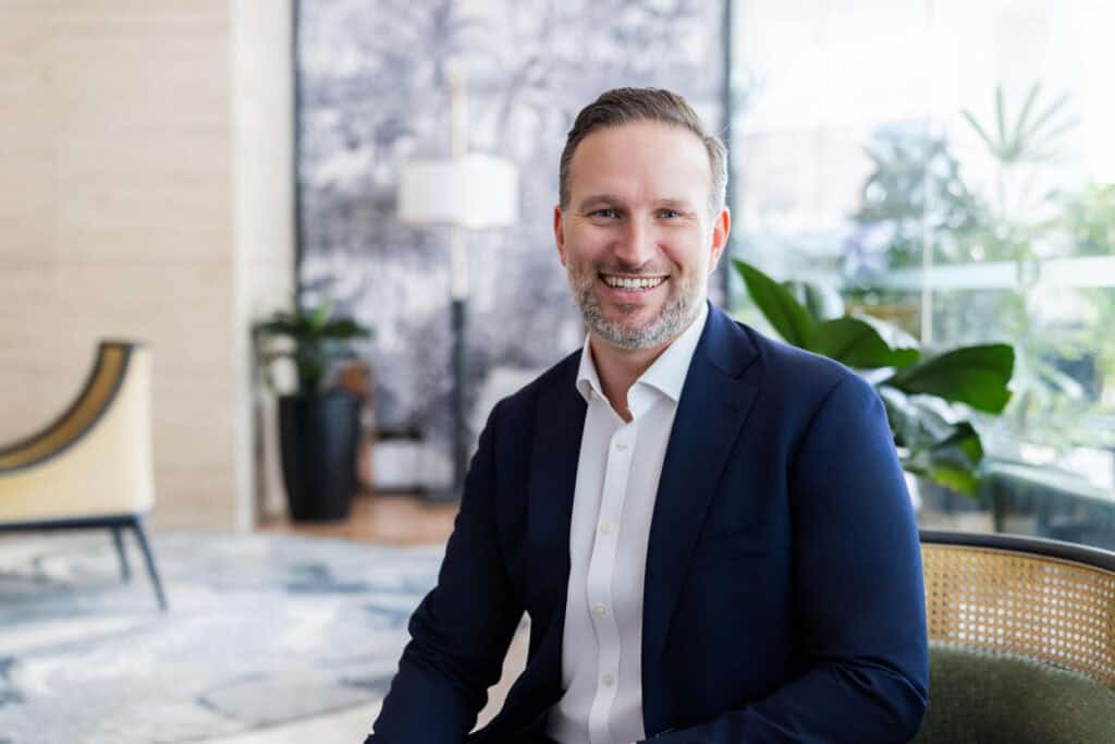 A man with a beard, wearing a blue suit and white shirt, smiling while seated in a modern lobby.