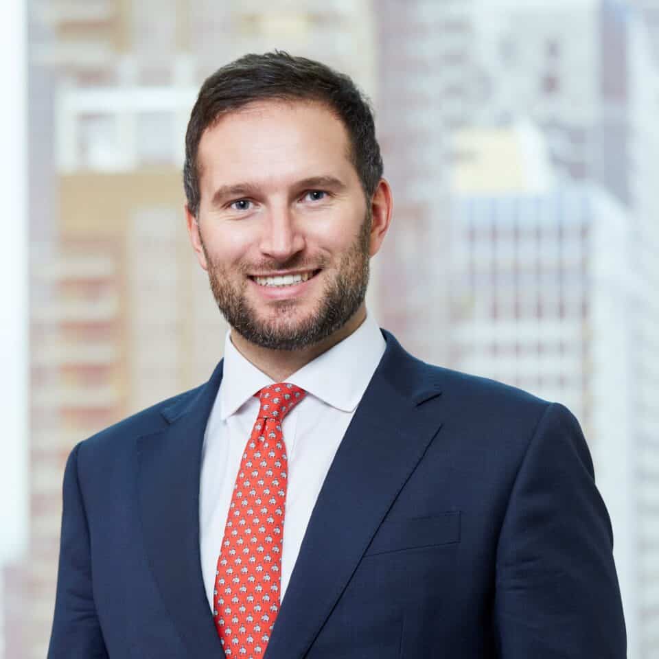 Headshot of a smiling man in a dark blue suit and a red tie with elephant patterns, against a blurred city background.