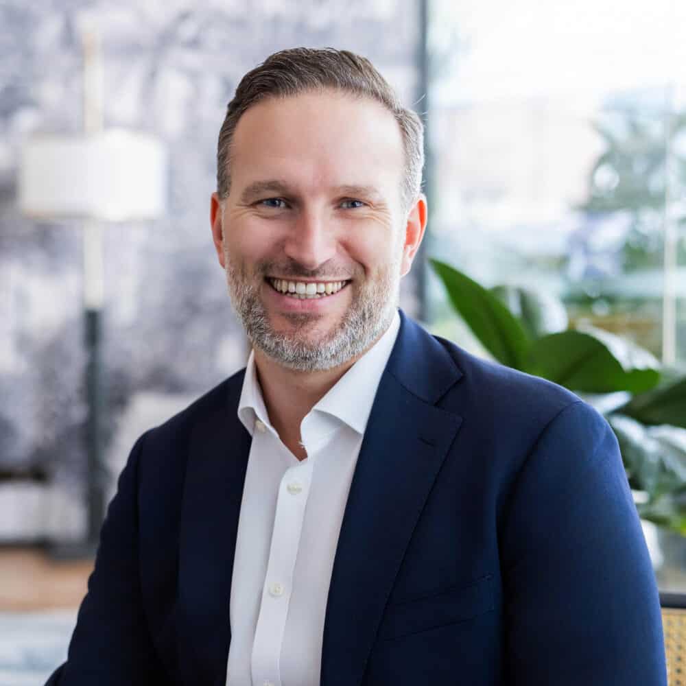 A man with a beard, wearing a blue suit and white shirt, smiling while seated in a modern lobby.