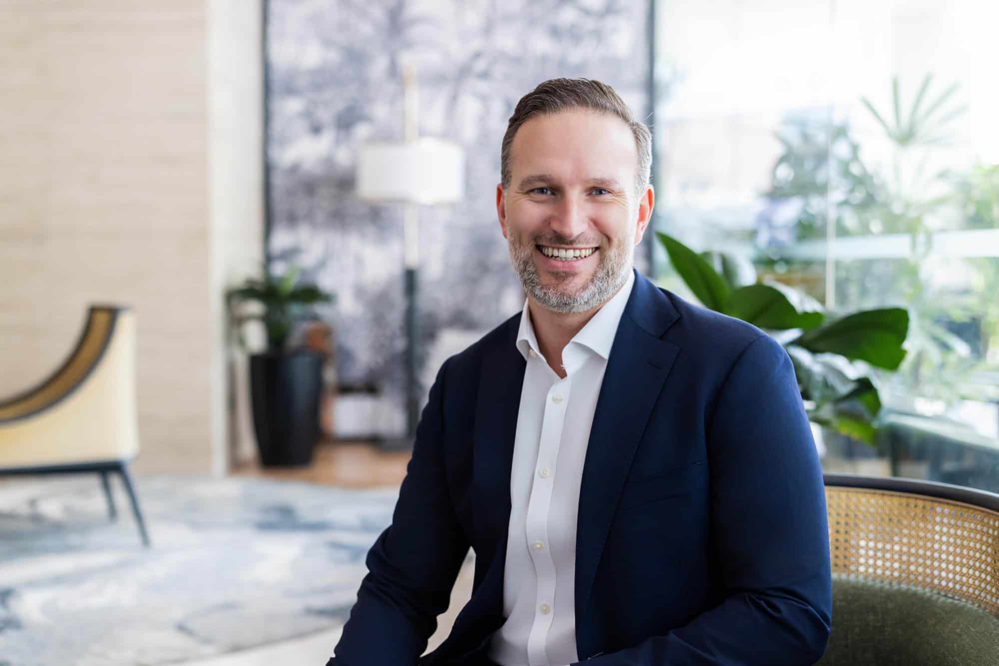 A man with a beard, wearing a blue suit and white shirt, smiling while seated in a modern lobby.